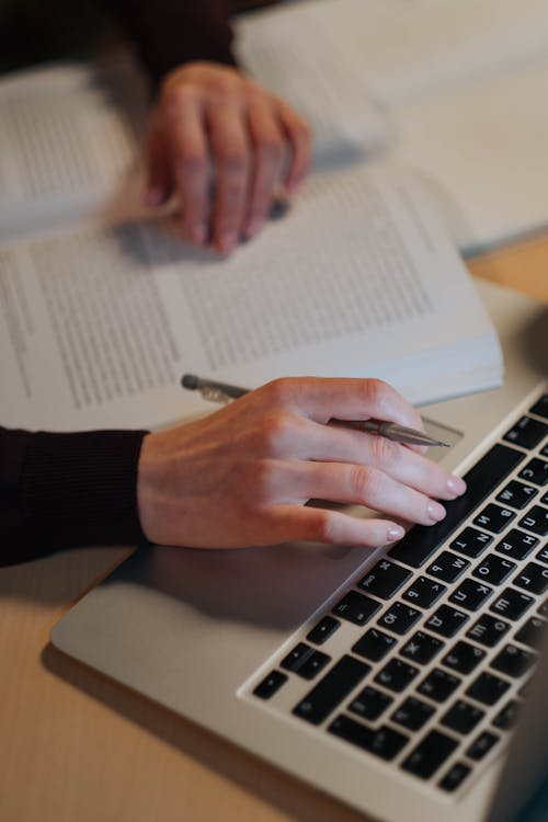 Graduate student using a laptop while holding a pen with a book open beside them during thesis writing and research planning.